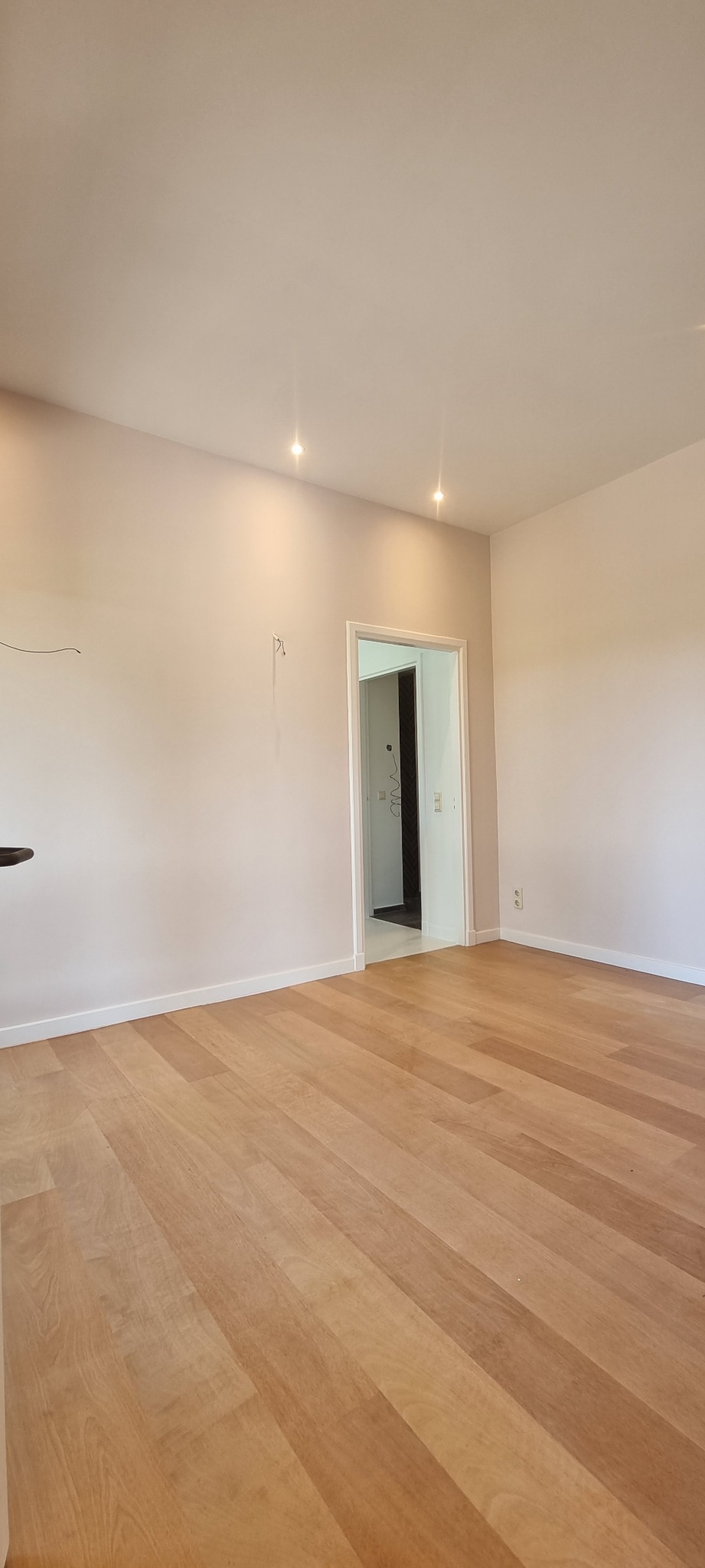 Pusty pokój z drewnianą podłogą, świeżo pomalowanymi ścianami w odcieniach beżu i dwiema lampami sufitowymi, widok na otwarte drzwi do kolejnego pomieszczenia.