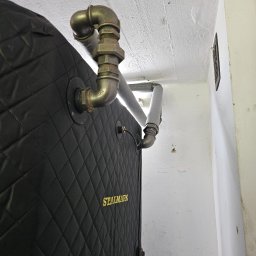 EKOTERM Andrzej Białoszewski - Częściowo widoczny piec z czarną, pikowaną obudową i napisem STALMARK, podłączony do metalowych rur instalacji grzewczej, biegnących wzdłuż białego sufitu i ściany.