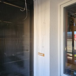 EKOTERM Andrzej Białoszewski - Ciemny kolektor słoneczny z wiszącymi przewodami montażowymi w trakcie instalacji wewnątrz pomieszczenia, widok z boku okna.