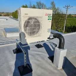 Biała jednostka zewnętrzna klimatyzacji Mitsubishi Electric zamontowana na dachu budynku, widoczne czarne gumowe podkładki antywibracyjne, rura odprowadzająca skropliny i elementy mocujące. W tle...