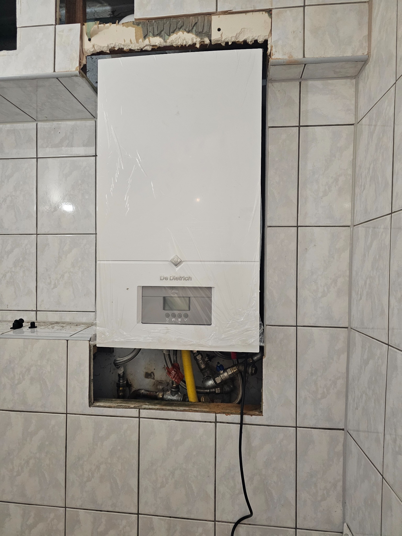 Nowy, biały piec gazowy marki De Dietrich, jeszcze w folii ochronnej, zamontowany w płytkach ceramicznych, z widocznymi rurami i przewodami w dolnej części.