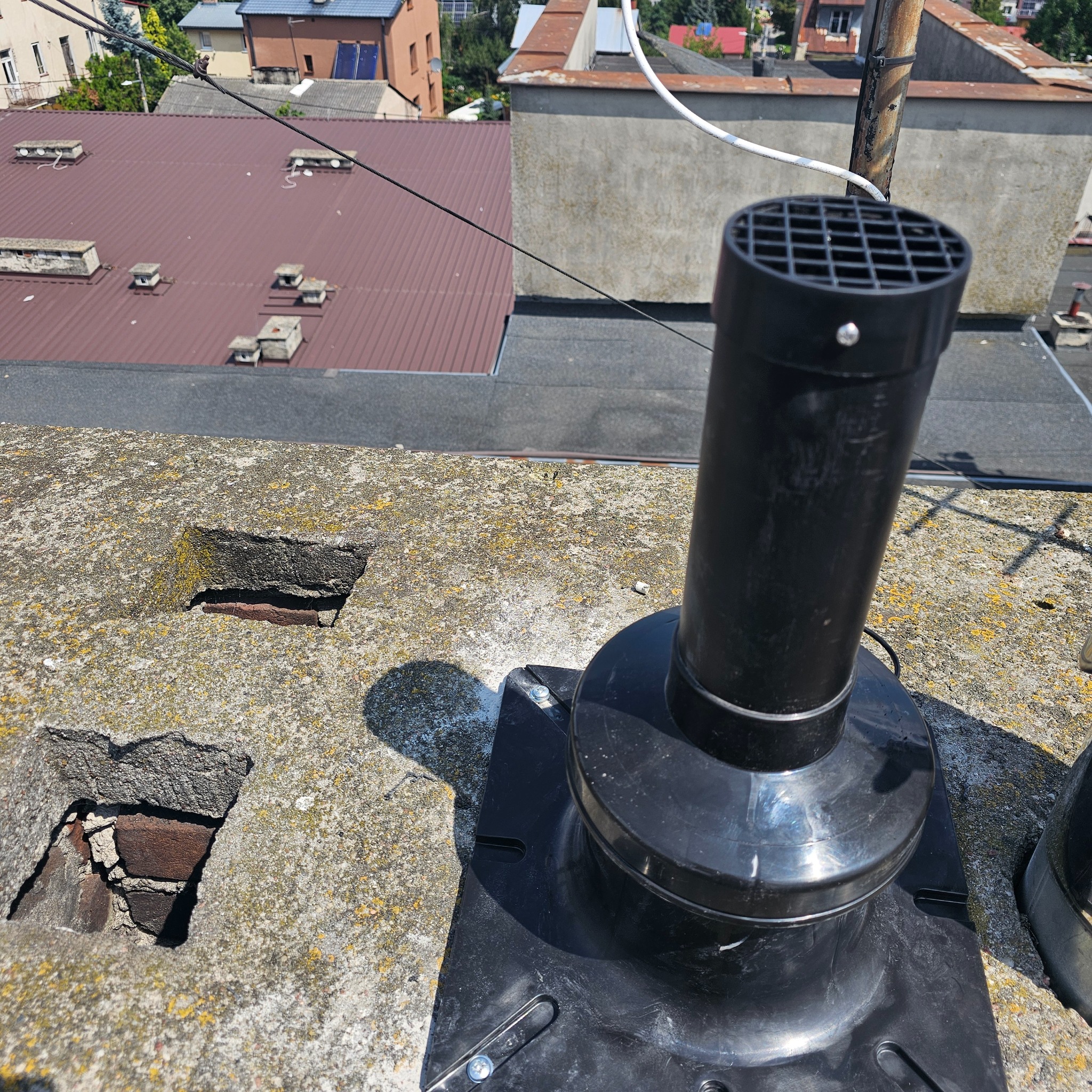 Czarny kominek wentylacyjny z kratką zamontowany na betonowym dachu, widoczne uszkodzenia w betonie i fragmenty cegieł.