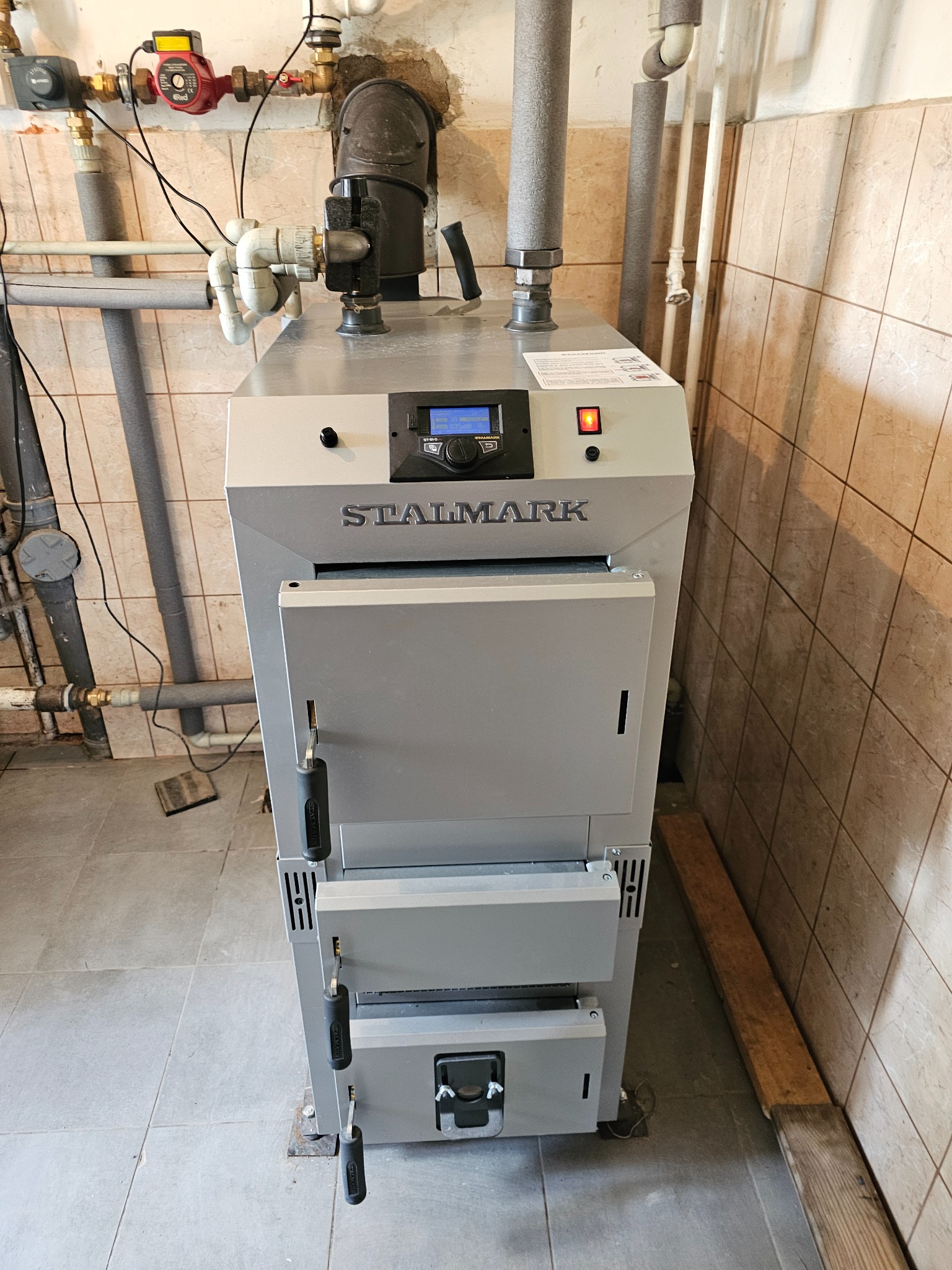 Szary piec CO marki Stalmark z elektronicznym wyświetlaczem i czerwonym wskaźnikiem LED, widoczne rury instalacji grzewczej i płytki ceramiczne na ścianie.