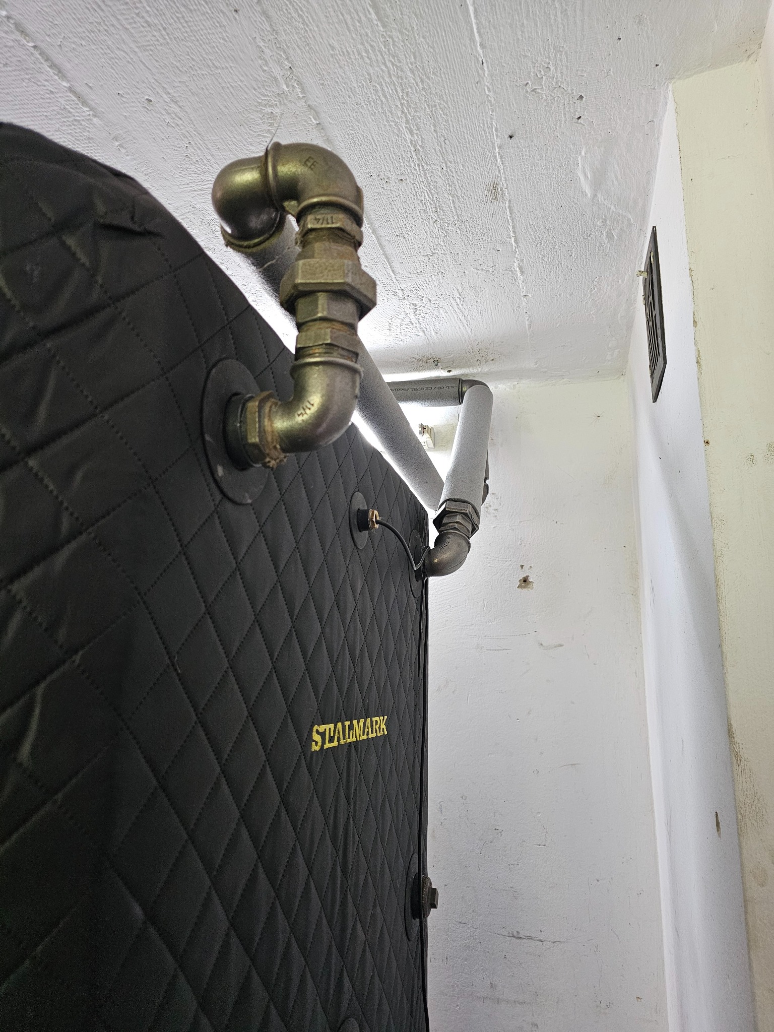 Częściowo widoczny piec z czarną, pikowaną obudową i napisem STALMARK, podłączony do metalowych rur instalacji grzewczej, biegnących wzdłuż białego sufitu i ściany.