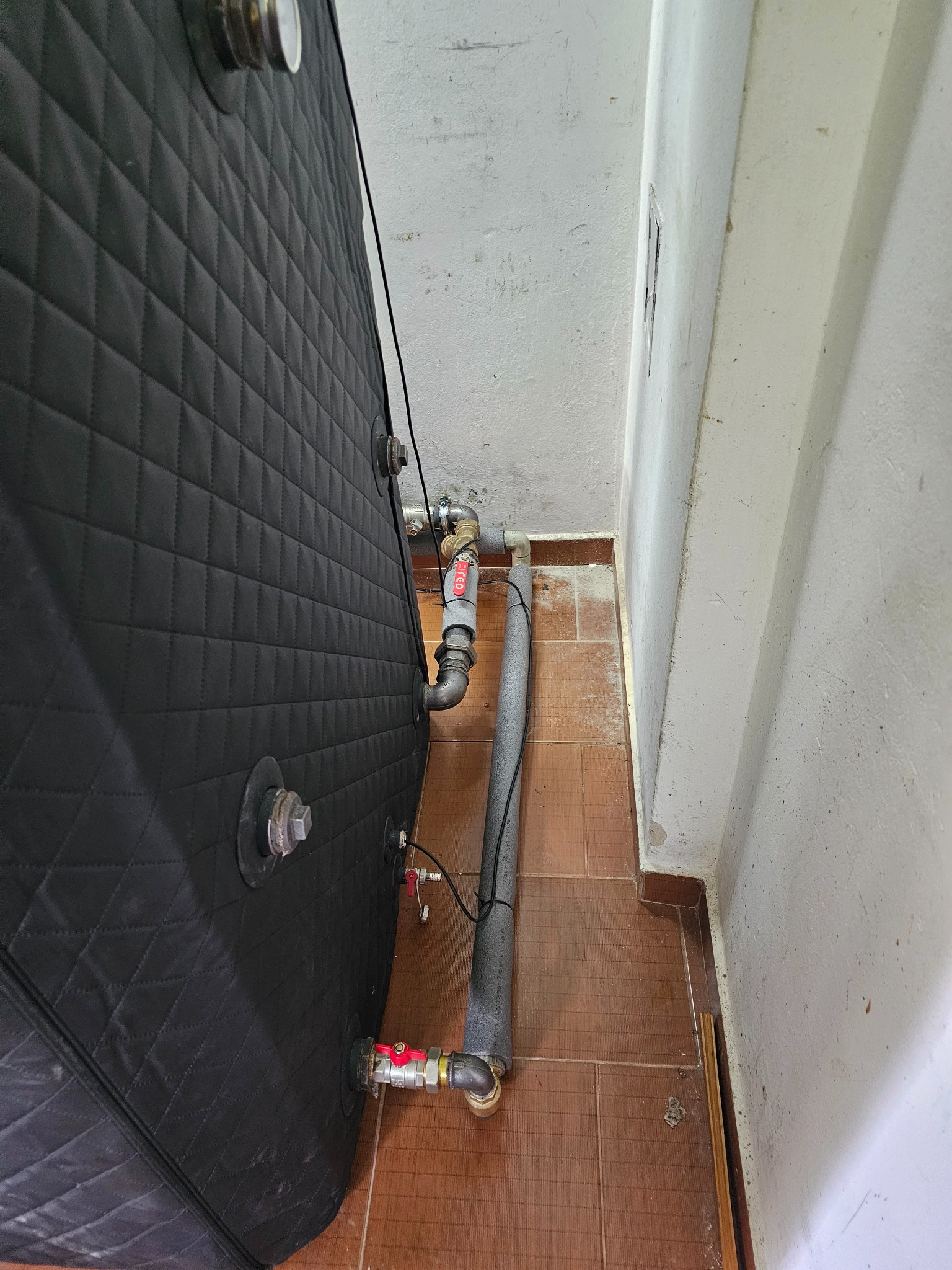 Czarny, ocieplony piec z widocznymi przyłączami rur i zaworami w pomieszczeniu z brudnymi ścianami i brązową podłogą z płytek.