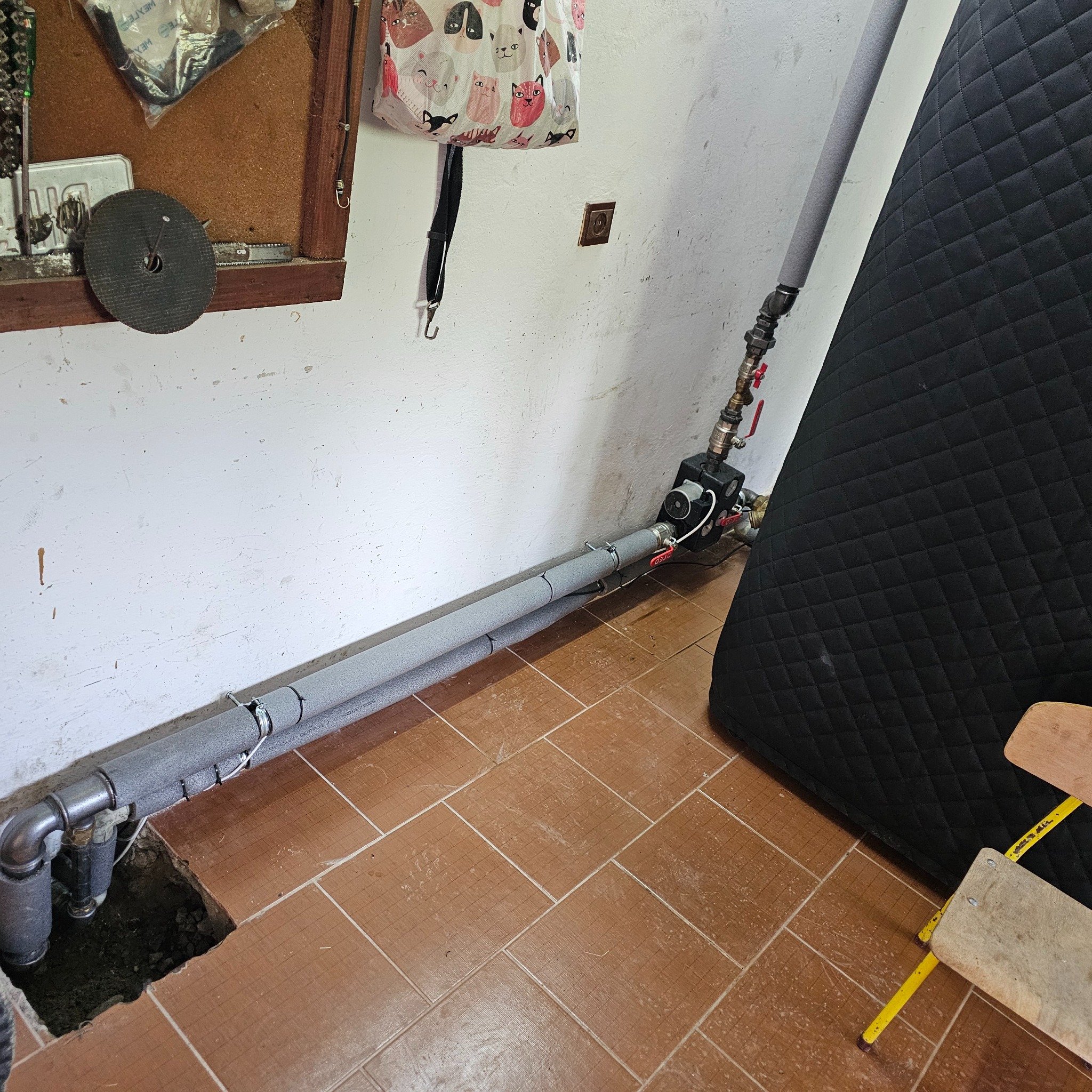 Fragment instalacji centralnego ogrzewania z zaizolowanymi rurami, pompą obiegową i czarnym zbiornikiem wyrównawczym w kotłowni, widoczne brązowe płytki podłogowe i otwór w podłodze z wystającymi...