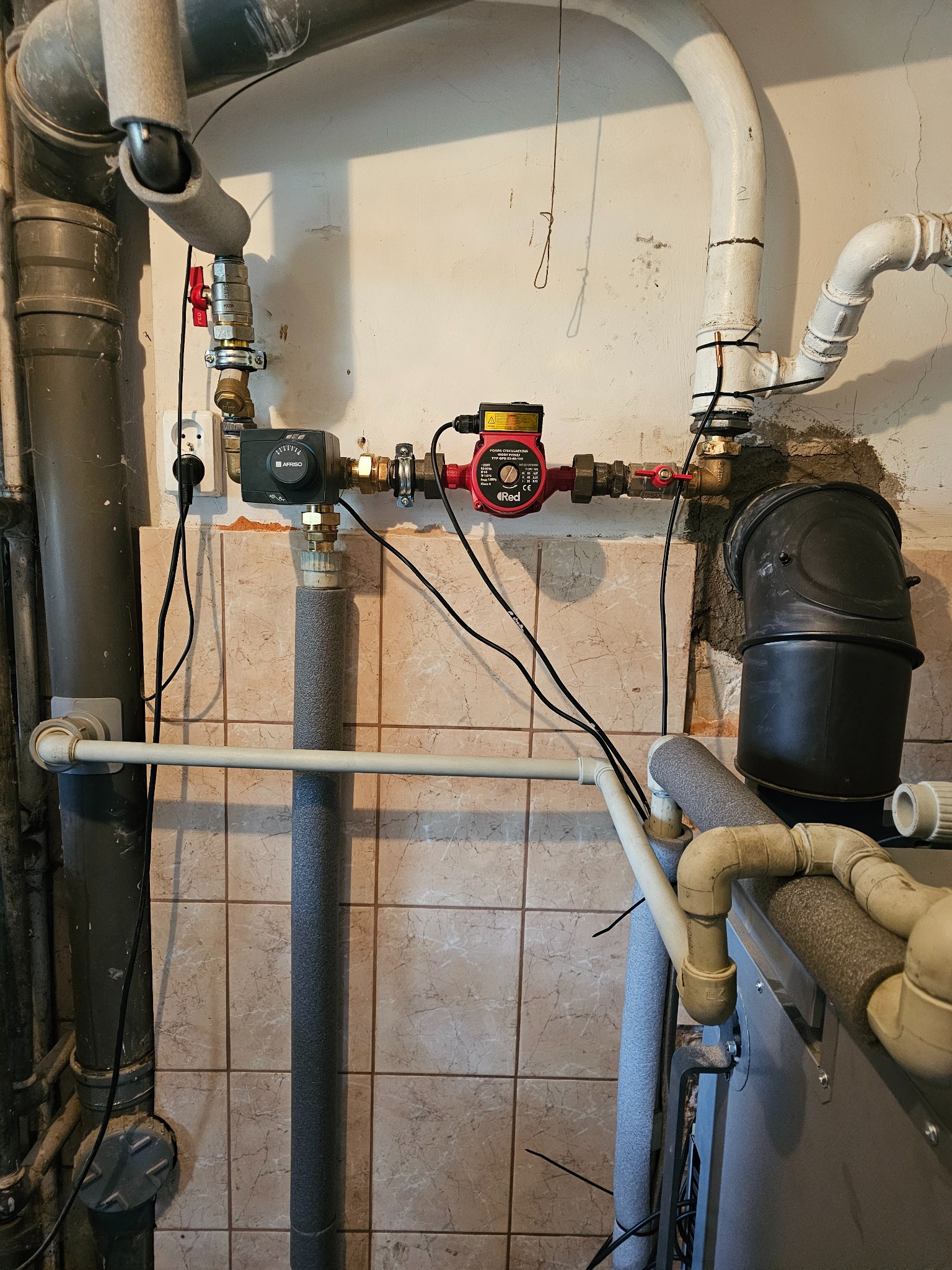 Fragment kotłowni z widocznym piecem, rurami grzewczymi z izolacją, pompą obiegową i zaworami na tle ściany wyłożonej kafelkami.