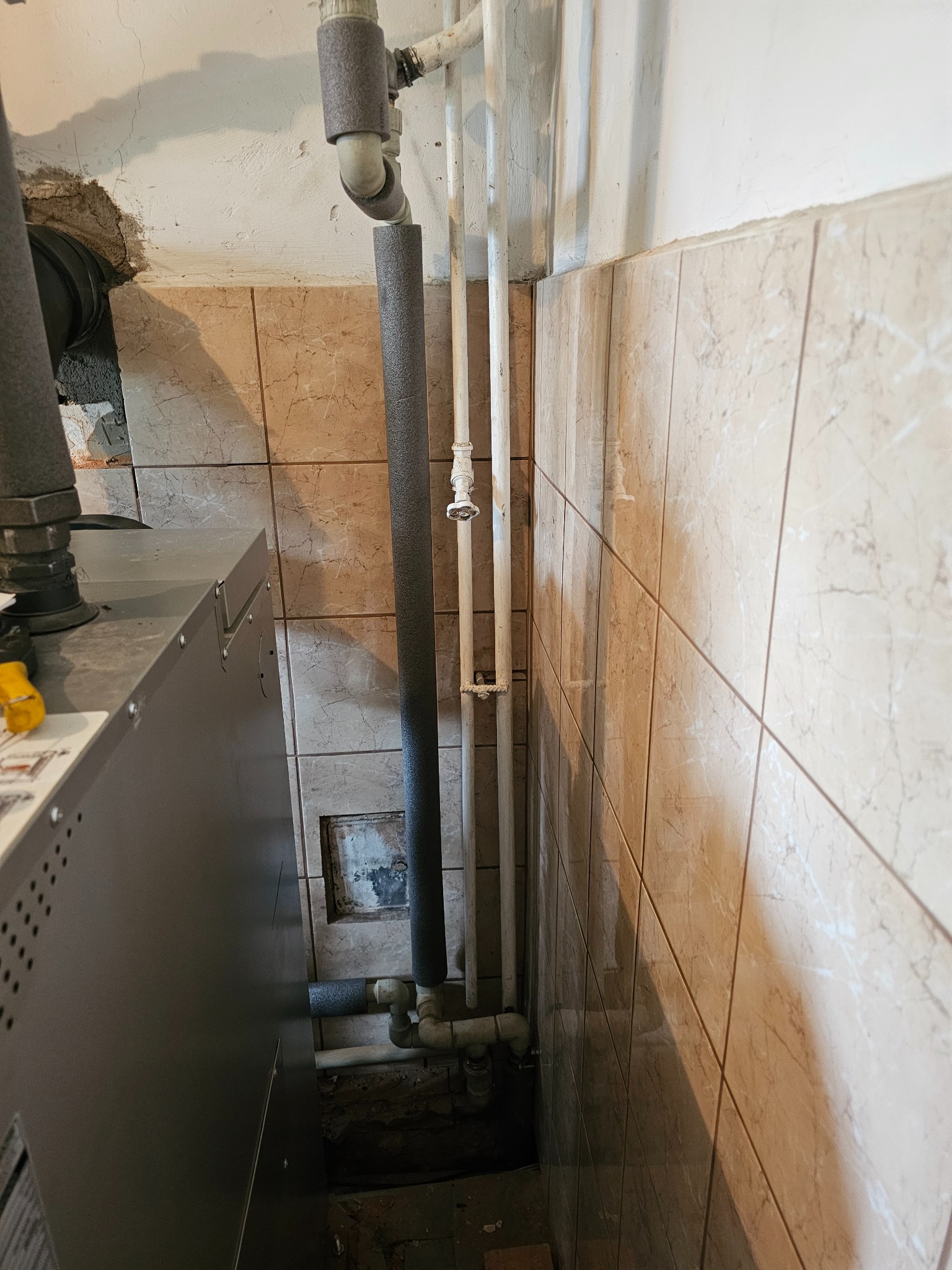 Szare rury grzewcze z izolacją termiczną prowadzone wzdłuż ściany wyłożonej beżowymi płytkami, widoczny fragment szarego pieca, podłoga z zabrudzeniami budowlanymi.
