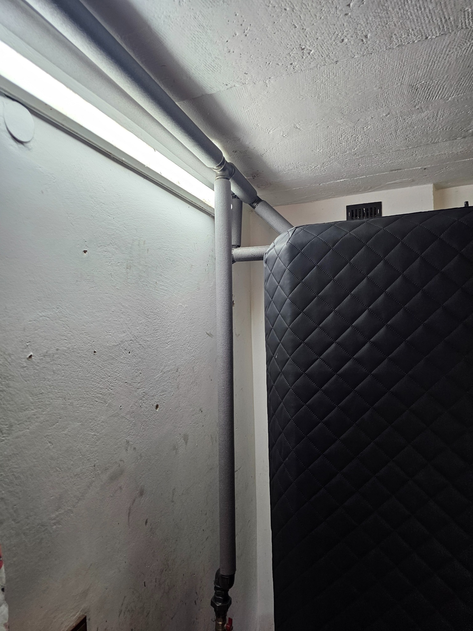 Szare, zaizolowane rury grzewcze biegnące wzdłuż ściany i sufitu w piwnicy, z czarnym, pikowanym zbiornikiem buforowym w tle.