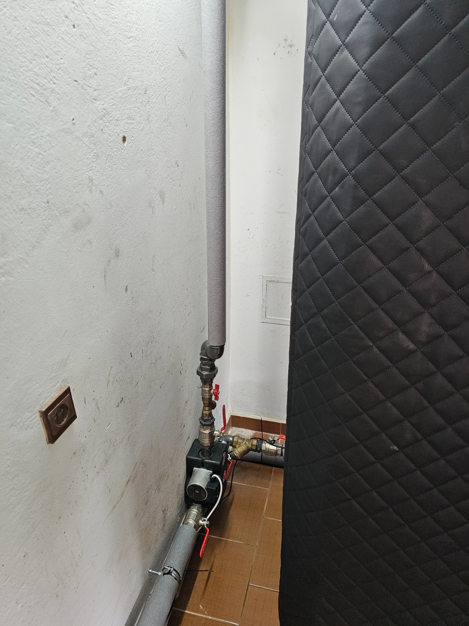 Fragment instalacji grzewczej z pompą obiegową, rurami w szarej izolacji i zaworami z czerwonymi uchwytami, widoczna na tle białej, zabrudzonej ściany i czarnego, pikowanego panelu.