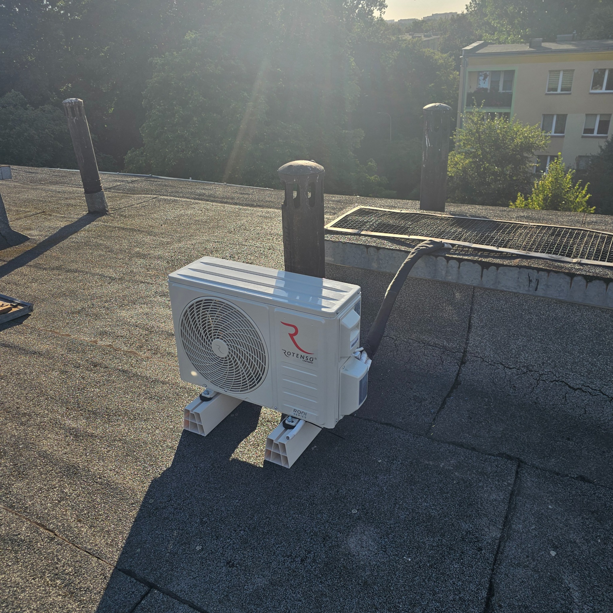 Biała jednostka zewnętrzna klimatyzacji Rotenso serii Roni zamontowana na dachu budynku z widocznymi kominami wentylacyjnymi i budynkami w tle.