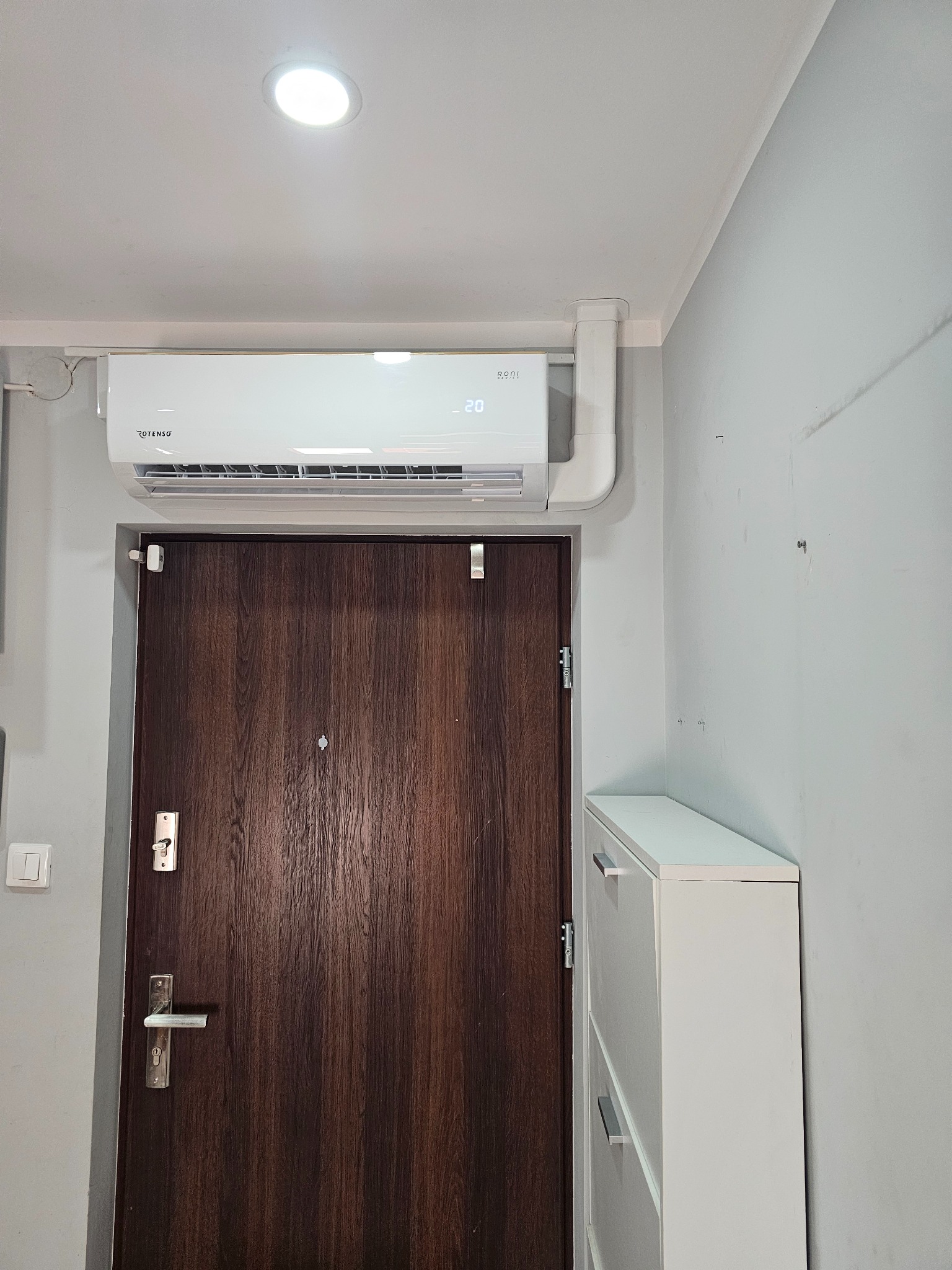 Biała jednostka klimatyzacji ROTENSO zamontowana nad ciemnobrązowymi drzwiami wejściowymi w jasnym korytarzu, widoczna biała szafka na buty.