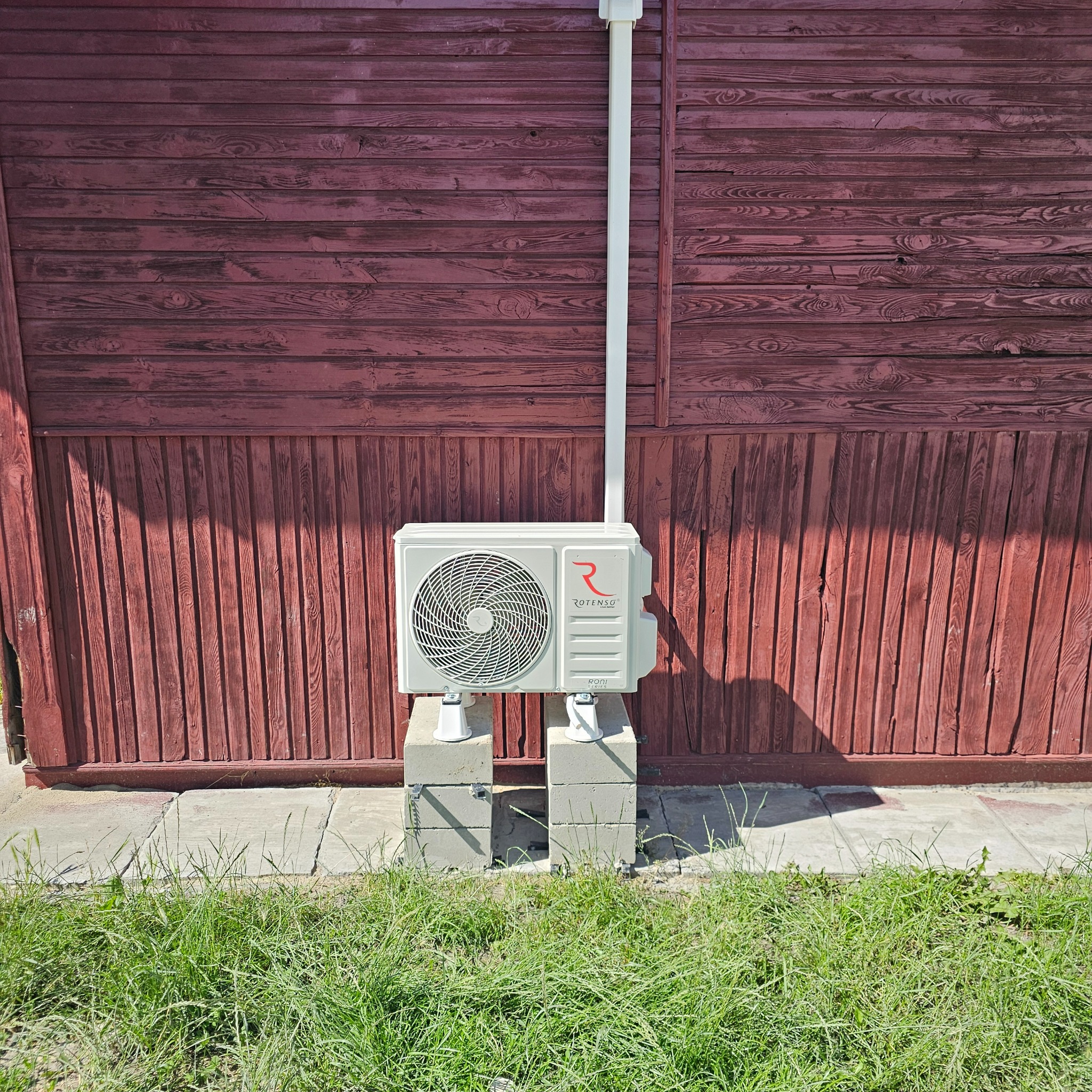 Biała jednostka zewnętrzna klimatyzatora Rotenso Roni Series zamontowana na betonowych bloczkach przed czerwoną, drewnianą ścianą budynku, z widoczną rurą odprowadzającą skropliny i fragmentem...