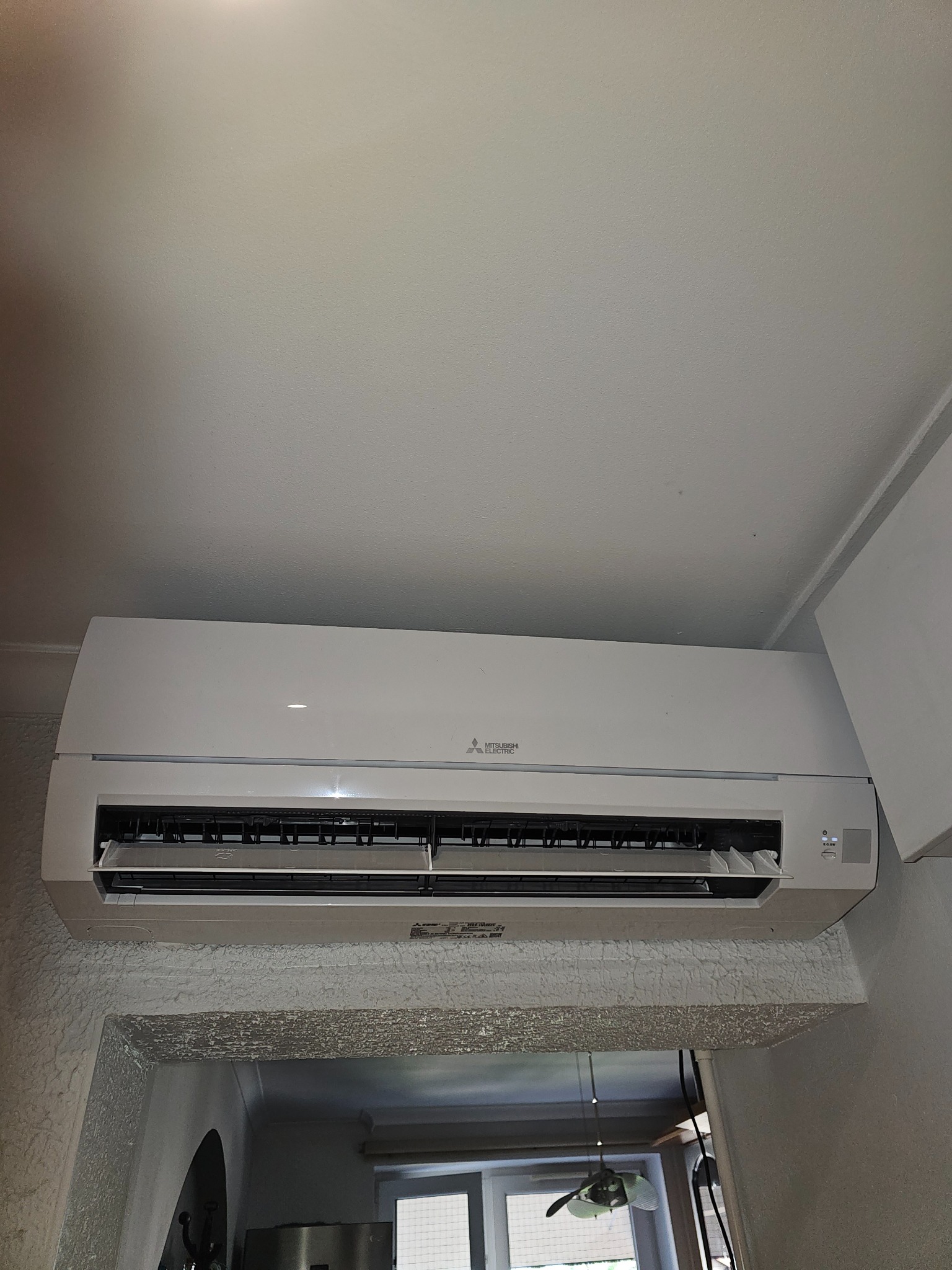 Biała jednostka wewnętrzna klimatyzacji Mitsubishi Electric zamontowana nad przejściem w pomieszczeniu z teksturowaną ścianą.