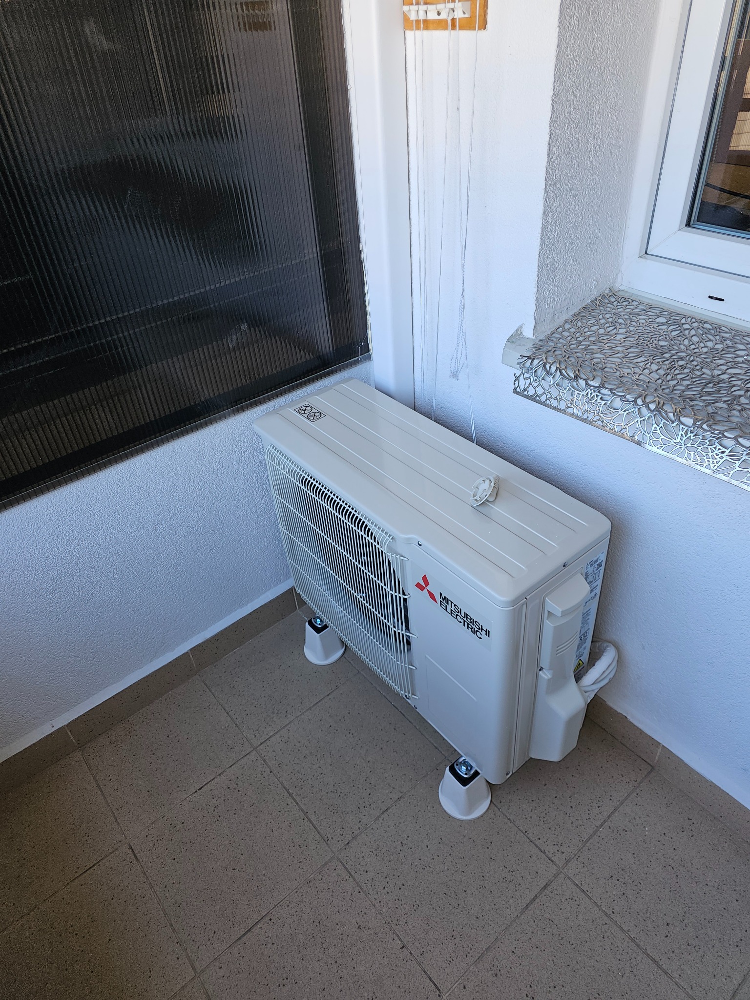 Biała jednostka zewnętrzna klimatyzacji Mitsubishi Electric umieszczona na czterech podkładkach, stoi na brązowych płytkach w rogu balkonu, widoczna czarna żaluzja okienna.