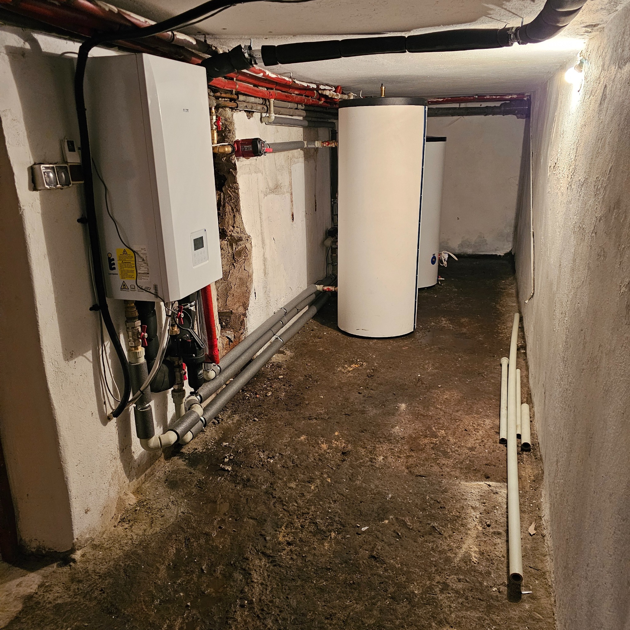 Piwnica z zainstalowanym piecem gazowym, zbiornikiem na wodę i rurami grzewczymi; widoczne niedoskonałości ścian i podłogi.