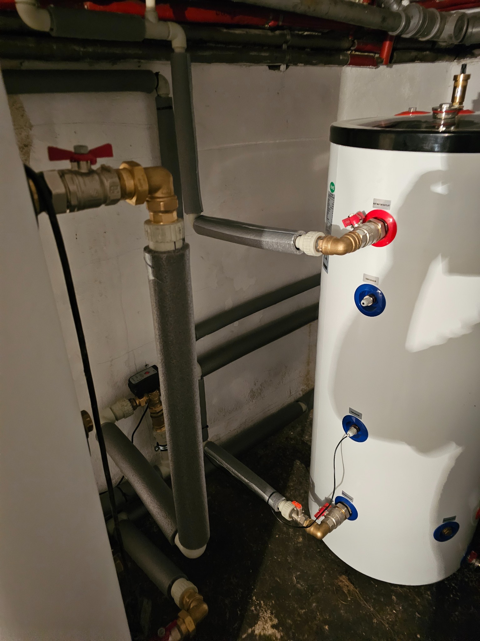 Biały zasobnik ciepłej wody użytkowej z podłączeniami hydraulicznymi, zaworami z czerwonymi uchwytami i czujnikami temperatury w niebieskich gniazdach, widoczne zaizolowane rury.