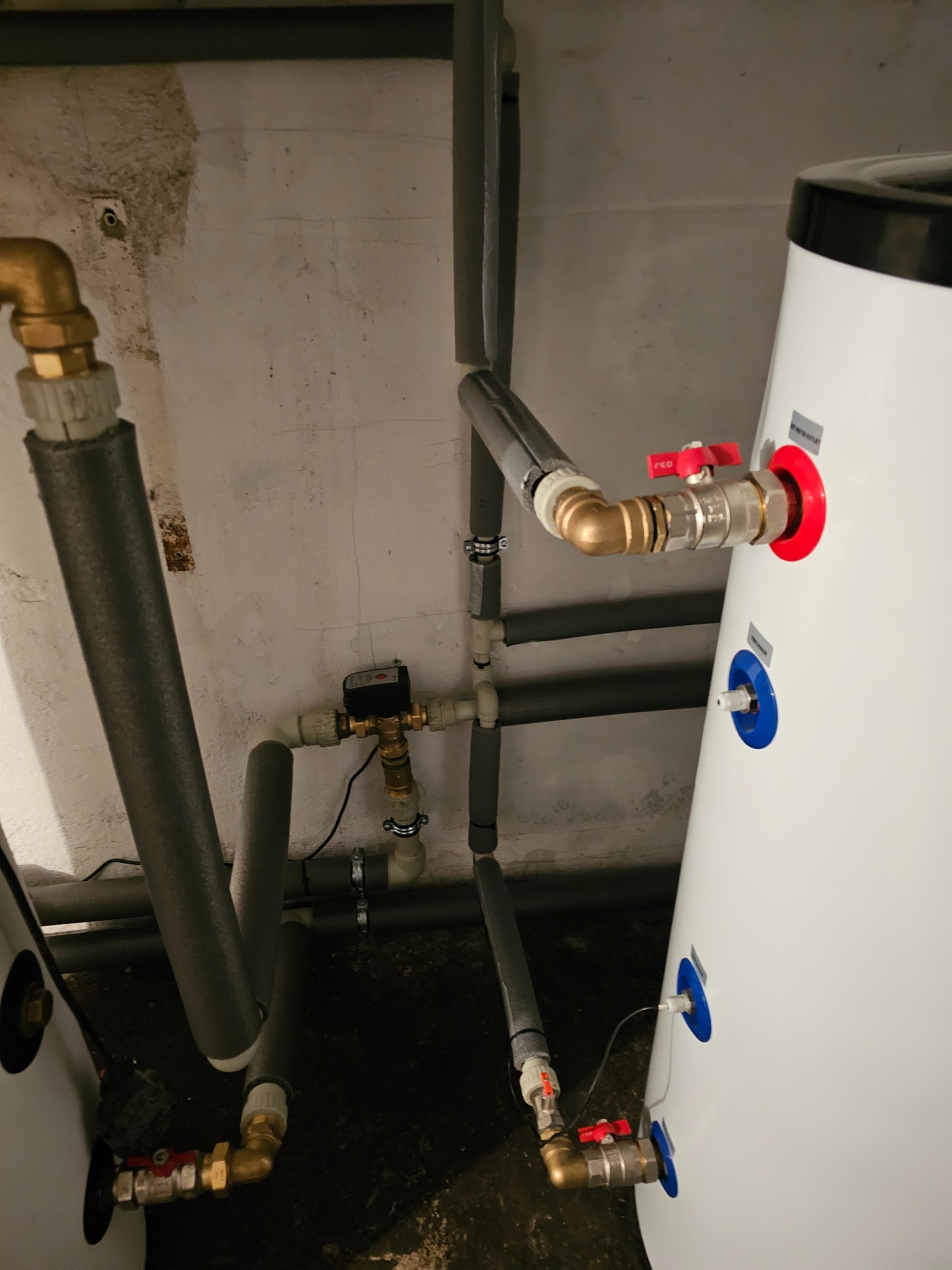 Biały zasobnik ciepłej wody użytkowej z podłączonymi rurami grzewczymi, zaworami kulowymi z czerwonymi uchwytami i czujnikiem temperatury, instalacja w surowym pomieszczeniu.