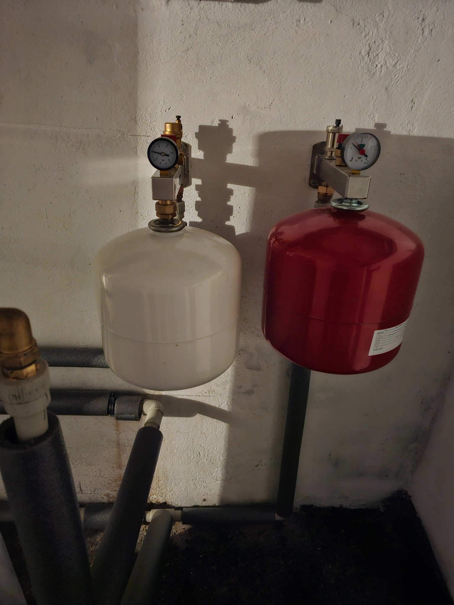 Dwa zbiorniki wyrównawcze ciśnienia, biały i czerwony, zamontowane na ścianie w piwnicy, z widocznymi manometrami i rurami instalacji grzewczej z izolacją.