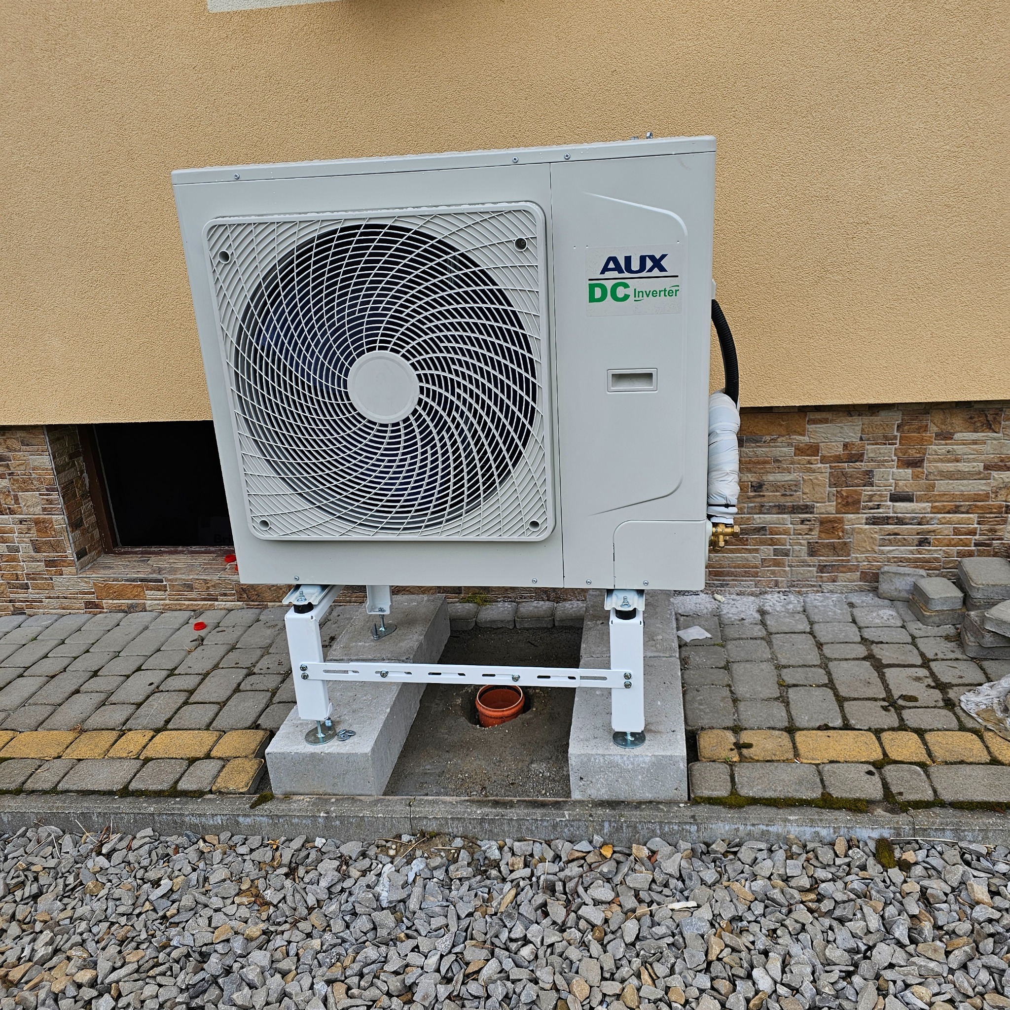 Zewnętrzna jednostka klimatyzacji AUX DC Inverter zamontowana na betonowym fundamencie i metalowych wspornikach przed budynkiem z jasną elewacją i kamienną podmurówką.