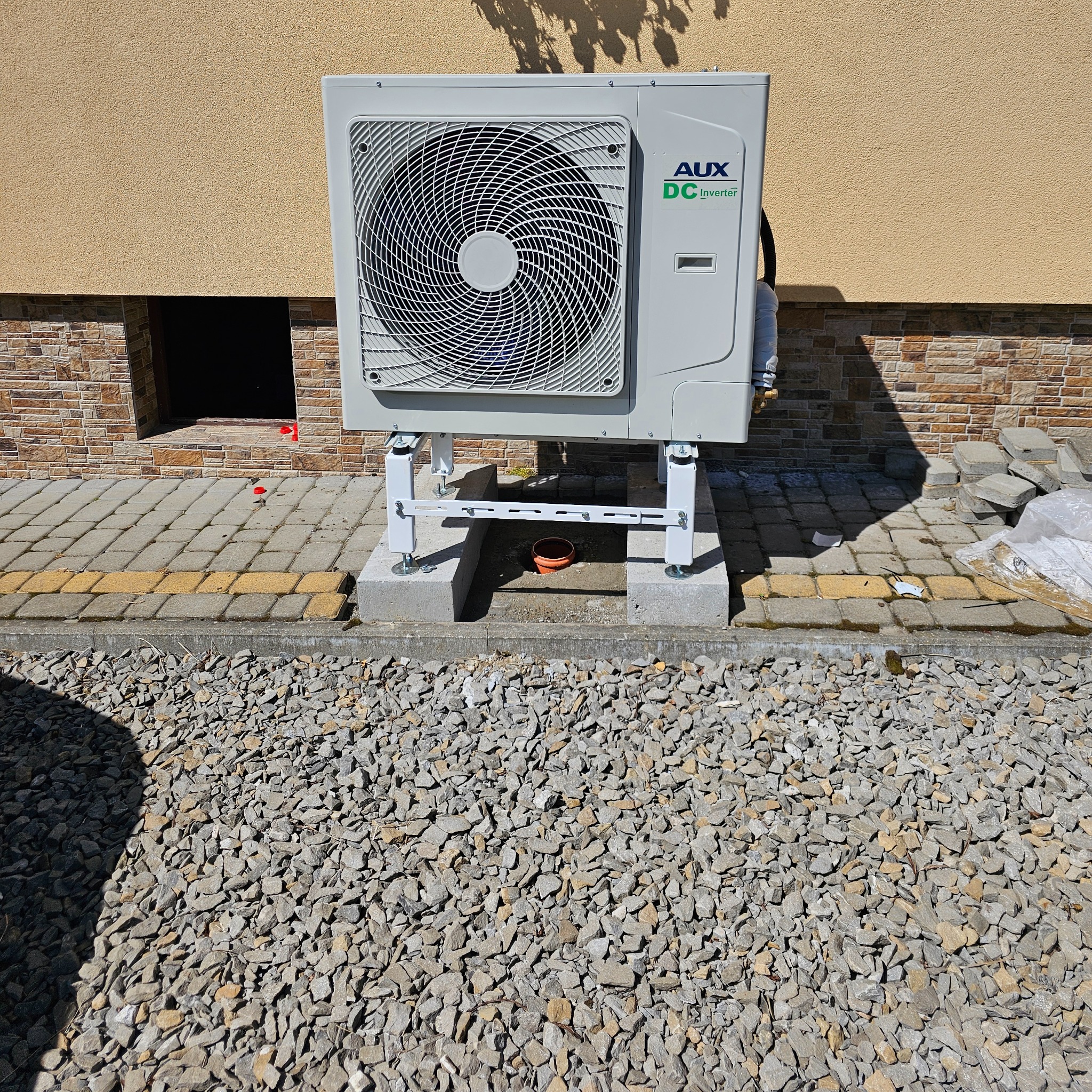 Zewnętrzna jednostka klimatyzacji AUX DC Inverter zamontowana na betonowych podstawach z metalowym stelażem, widoczne kamienne podłoże i fragment elewacji budynku z ceglaną podmurówką.
