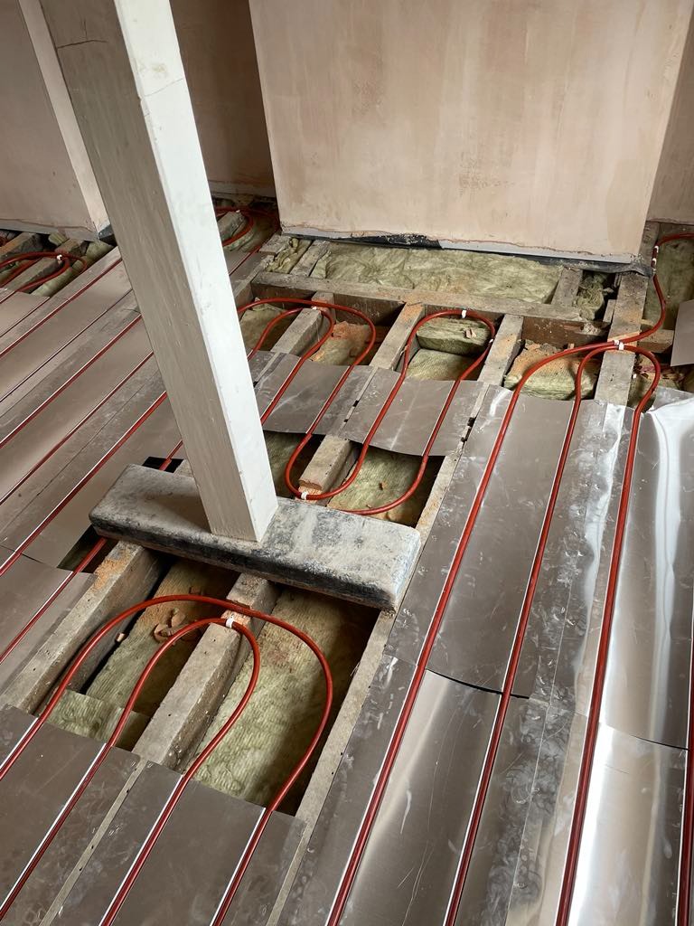 Montaż podłogowego ogrzewania wodnego z czerwonymi rurami i aluminiowymi płytami rozpraszającymi ciepło, widoczne ocieplenie między legarami podłogi.