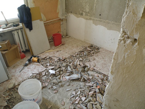 Demontaż starej podłogi z płytek przy użyciu młota udarowego. Widoczne gruz, fragmenty płytek, wiaderka i przygotowane ściany do dalszych prac remontowych.