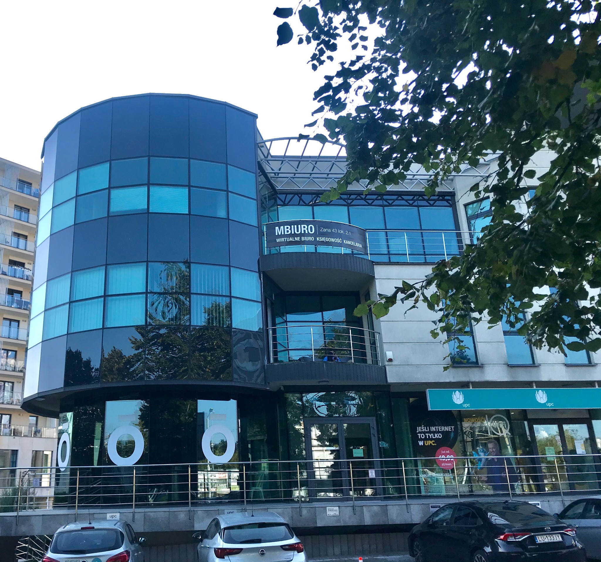 Nowoczesny budynek biurowy z zaokrągloną przeszkloną fasadą, na której widnieją duże białe cyfry, z balkonem, na którym umieszczono baner z napisem 'MBIURO wirtualne biuro księgowość kancelaria'...