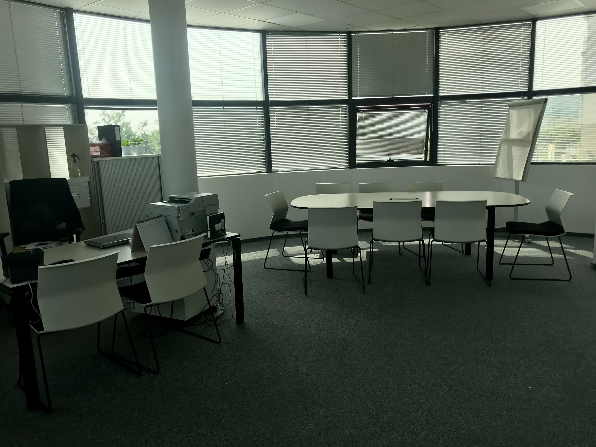 Jasne biuro z dużym oknem z żaluzjami, owalnym stołem konferencyjnym i białymi krzesłami, biurko z komputerem i drukarką z lewej strony, flipchart w rogu.