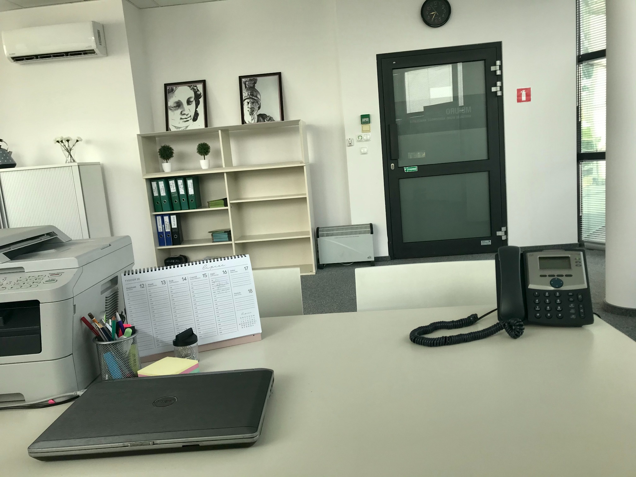 Wnętrze biura z biurkiem, na którym leży laptop, kalendarz biurkowy, telefon stacjonarny i przybory do pisania, w tle regał z segregatorami i dekoracjami.