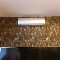 Zamontowany klimatyzator marki Gree na ścianie o dekoracyjnej fakturze we Wrocławiu, widoczny fragment telewizora i rośliny doniczkowej.