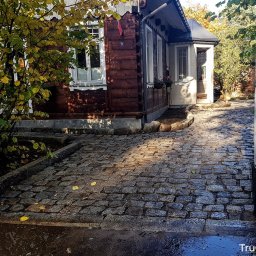 Trugarden Krzysztof Truszewski - Ułożona kostka granitowa na podjeździe do domu z drewnianą elewacją, widok zza metalowej bramy, jesienne liście na kostce.