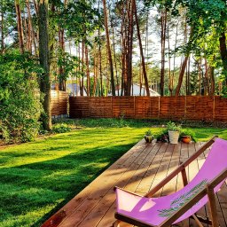 Trugarden Krzysztof Truszewski - Drewniany taras w ogrodzie z leżakiem w odcieniach różu, na tarasie donice z kwiatami, w tle trawnik i drewniany płot lamelowy