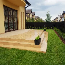 Drewnopodobny taras z trzema stopniami, z donicami na dwóch poziomach, sąsiadujący z trawnikiem i wysokim, ciemnym ogrodzeniem, na tle zabudowy jednorodzinnej w pochmurny dzień.