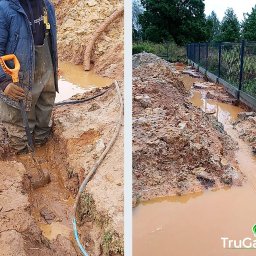 Trugarden Krzysztof Truszewski - Pracownik w wodoodpornych spodniobutach kopie rów w błotnistym terenie pod ogrodzeniem panelowym, używając szpadla z pomarańczowym uchwytem; obok widoczny kabel i rura.