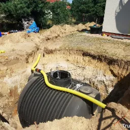 Czarny zbiornik zakopany w wykopie z żółtymi rurami podłączonymi do niego, widoczne narzędzia i fragment domu w tle.