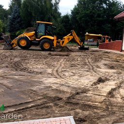 Trugarden Krzysztof Truszewski - Żółta koparko-ładowarka przygotowuje teren pod budowę kostki brukowej na posesji z ceglanym ogrodzeniem i domem w tle.