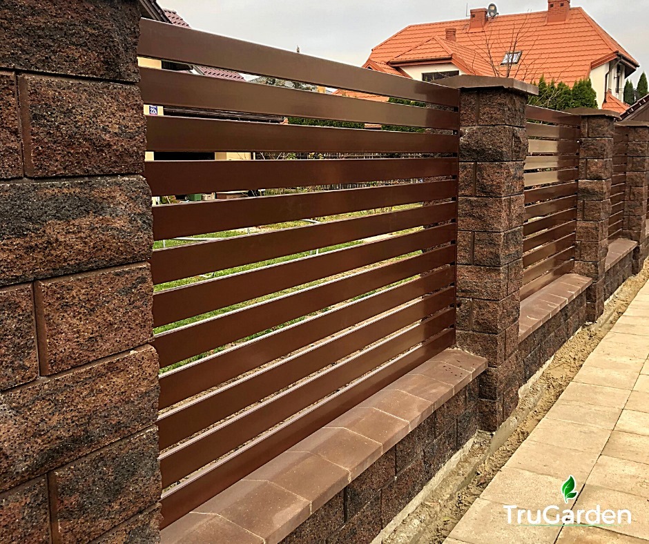 Nowoczesne ogrodzenie panelowe z brązowymi, poziomymi przęsłami i słupkami z brązowego kamienia łupanego, widok z boku na tle domu z czerwoną dachówką.