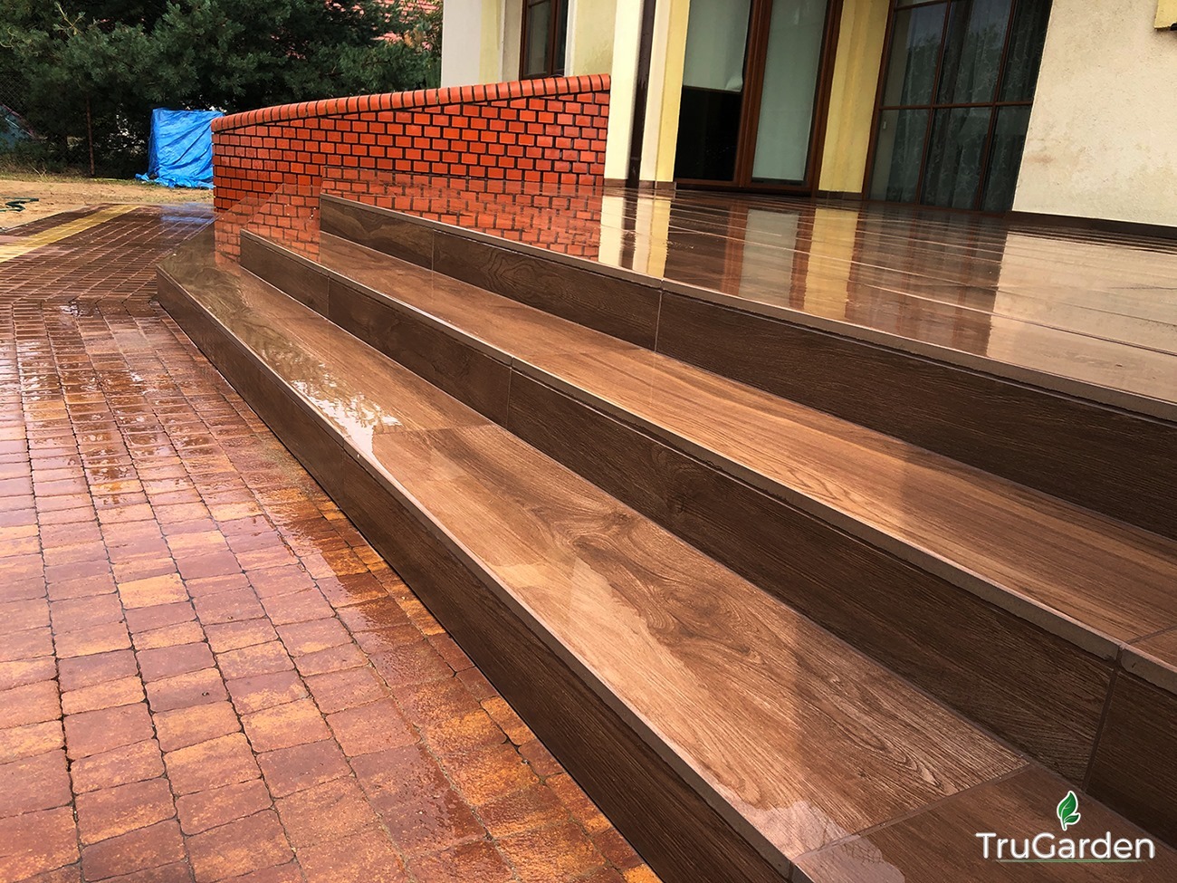Kaskada trzech stopni z płytek imitujących drewno, prowadząca z kostki brukowej do tarasu, po deszczu, widoczna ceglana ścianka i logo TruGarden w prawym dolnym rogu.