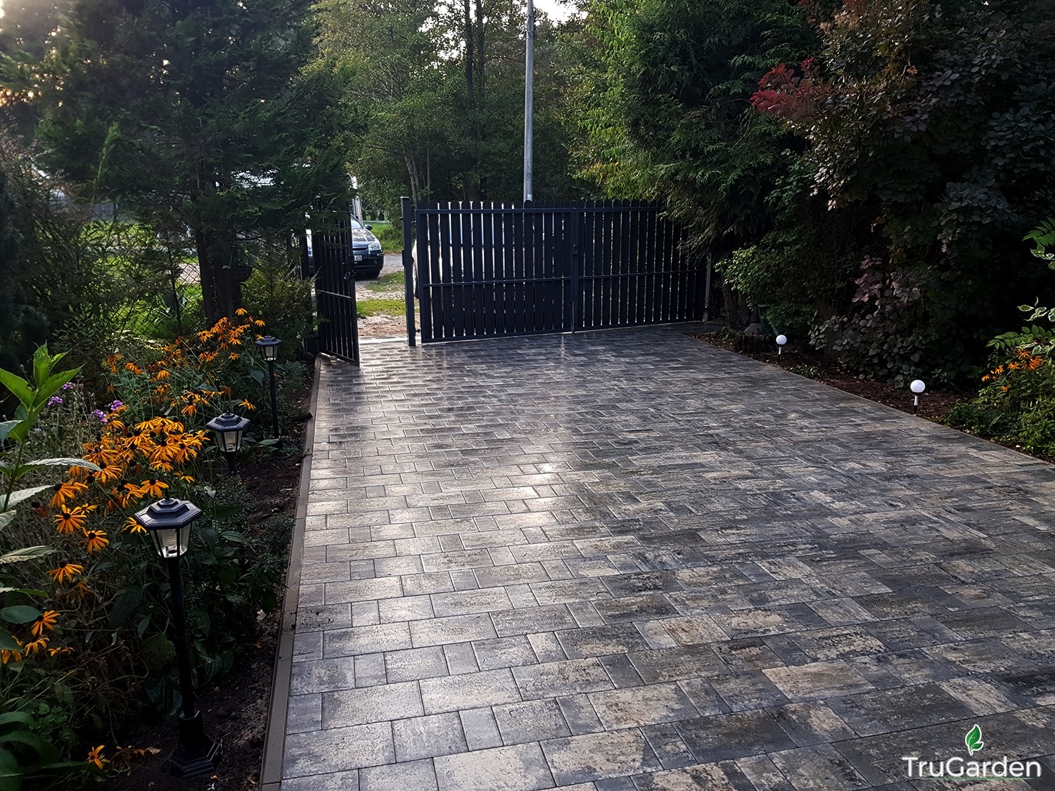Szeroki podjazd z szarej kostki brukowej, oświetlony lampami ogrodowymi, prowadzący do czarnej, metalowej bramy w otoczeniu zieleni i kwitnących roślin.