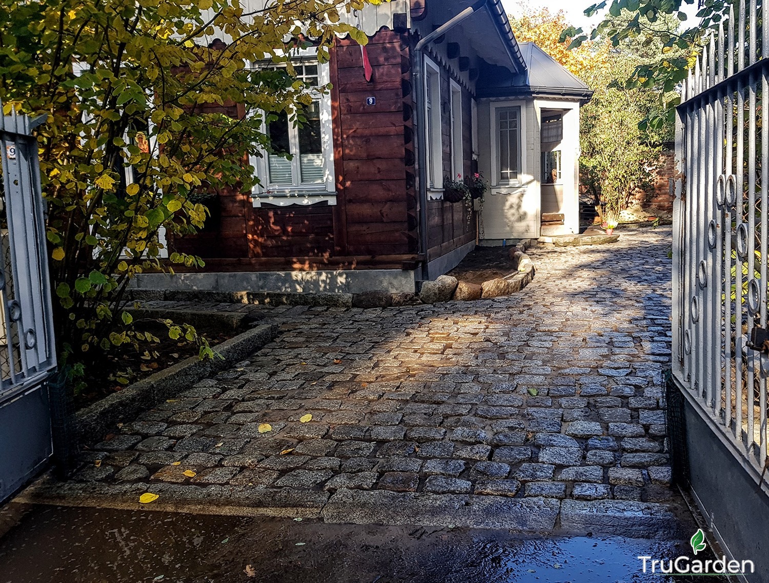 Ułożona kostka granitowa na podjeździe do domu z drewnianą elewacją, widok zza metalowej bramy, jesienne liście na kostce.