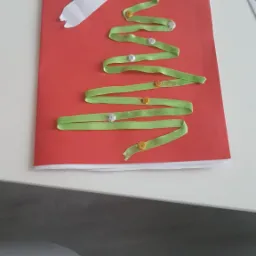 Ręcznie robiona kartka świąteczna z czerwonego papieru, ozdobiona zieloną wstążką ułożoną w kształt choinki z przyszytymi guzikami i białą wstążką na górze.