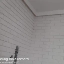 Pomieszczenie z wykończoną ścianą z białej cegły dekoracyjnej, widoczny narożnik i fragment sufitu z listwą przypodłogową, w dolnym rogu oparta drabina aluminiowa.