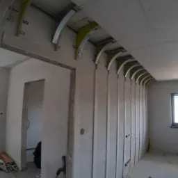 Konstrukcja sufitu podwieszanego z płyt kartonowo-gipsowych z elementami łukowymi, widoczne profile metalowe i drewniane wsporniki, instalacja elektryczna w ścianie.