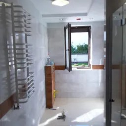 Wnętrze łazienki po remoncie z grzejnikiem drabinkowym, szafką pod umywalką z drewnianym akcentem, otwartym oknem na ogród i narzędziami do fugowania na podłodze.