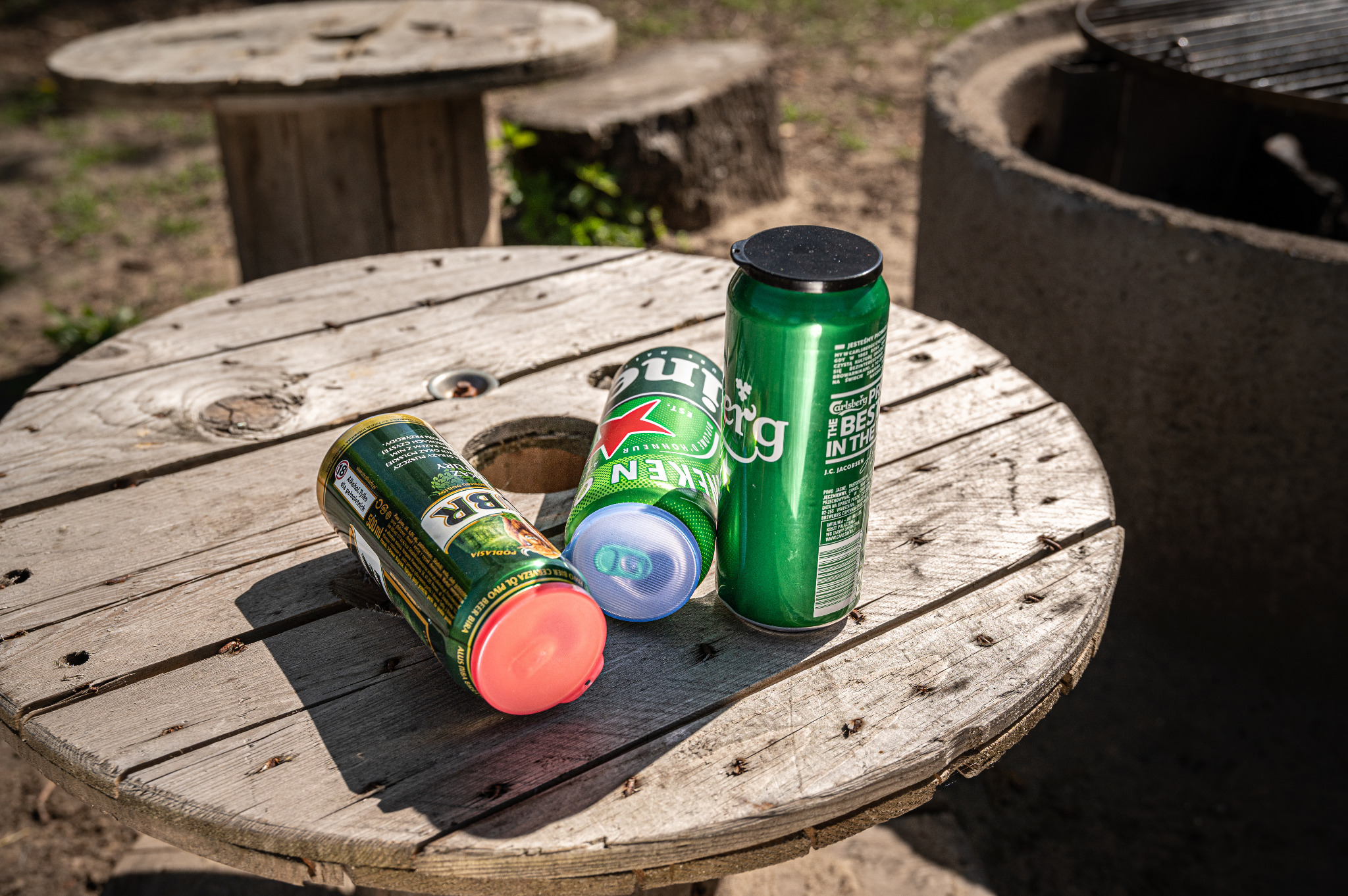 Trzy puszki piwa różnych marek (Carlsberg, Heineken, BR Podlasie) stojące na drewnianym stole zrobionym ze szpuli na kable, w tle betonowy grill.