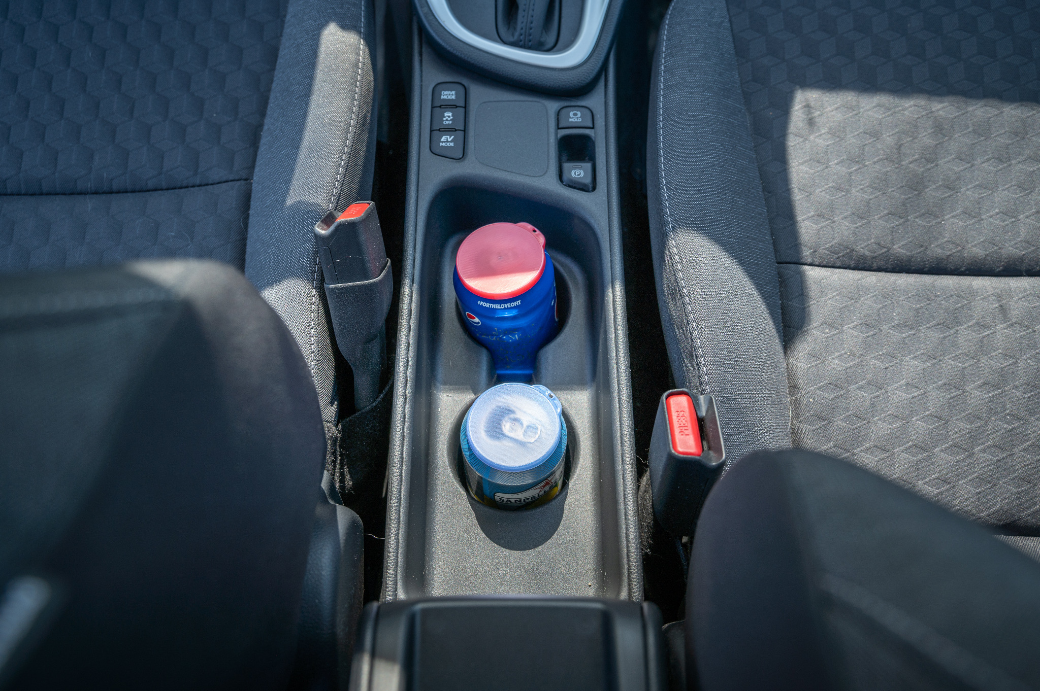 Dwa napoje w uchwycie samochodowym, jeden w puszce San Pellegrino i drugi w butelce Pepsi z różową zakrętką, widziane z góry w szarym wnętrzu auta.