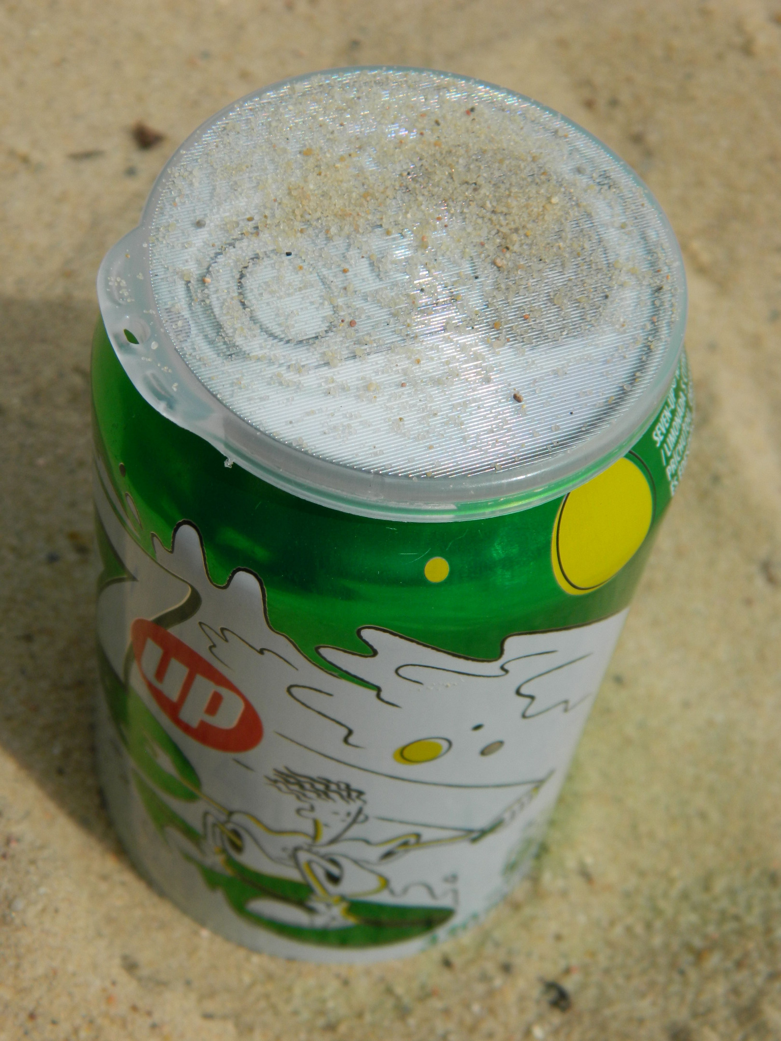 Otwarta puszka napoju 7up z plastikowym wieczkiem, częściowo zasypana piaskiem, stojąca na piasku.