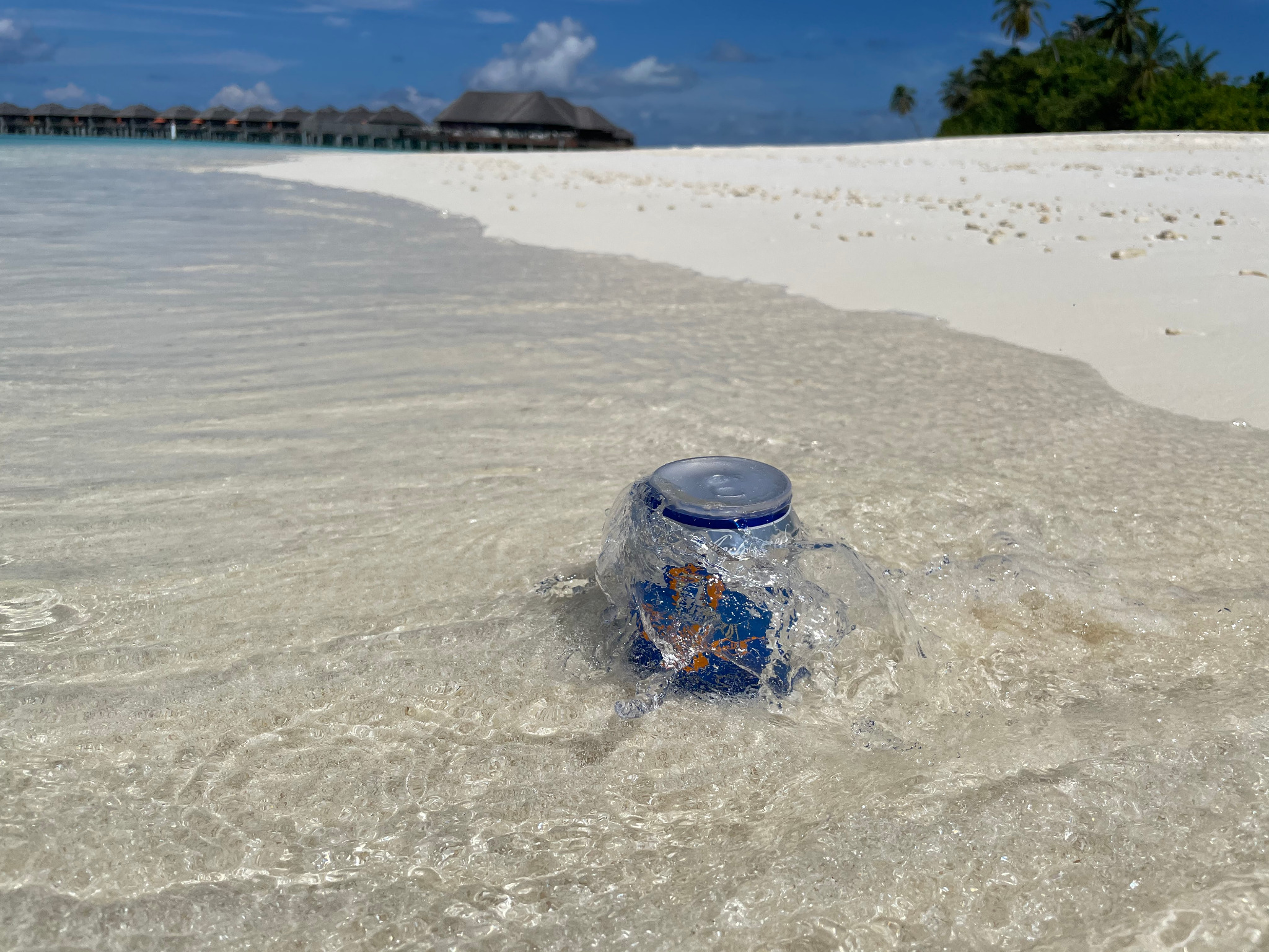 Niebieska puszka z wzorem w wodzie na piaszczystej plaży, w tle domki na wodzie i palmy.
