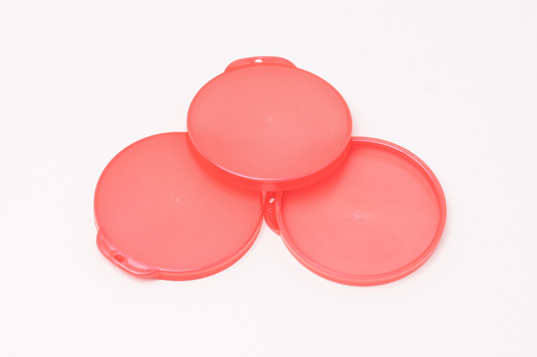 Trzy czerwone, okrągłe, plastikowe pokrywki z uchwytami ułożone jedna na drugiej na białym tle.